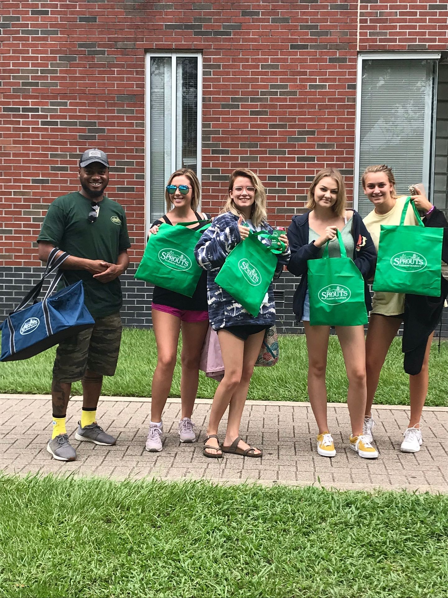 Group of people standing outside holding green Sprouts reusable bags in front of a brick building.