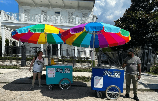 Two people standing beside ice cream carts with colorful umbrellas in front of a large white house.